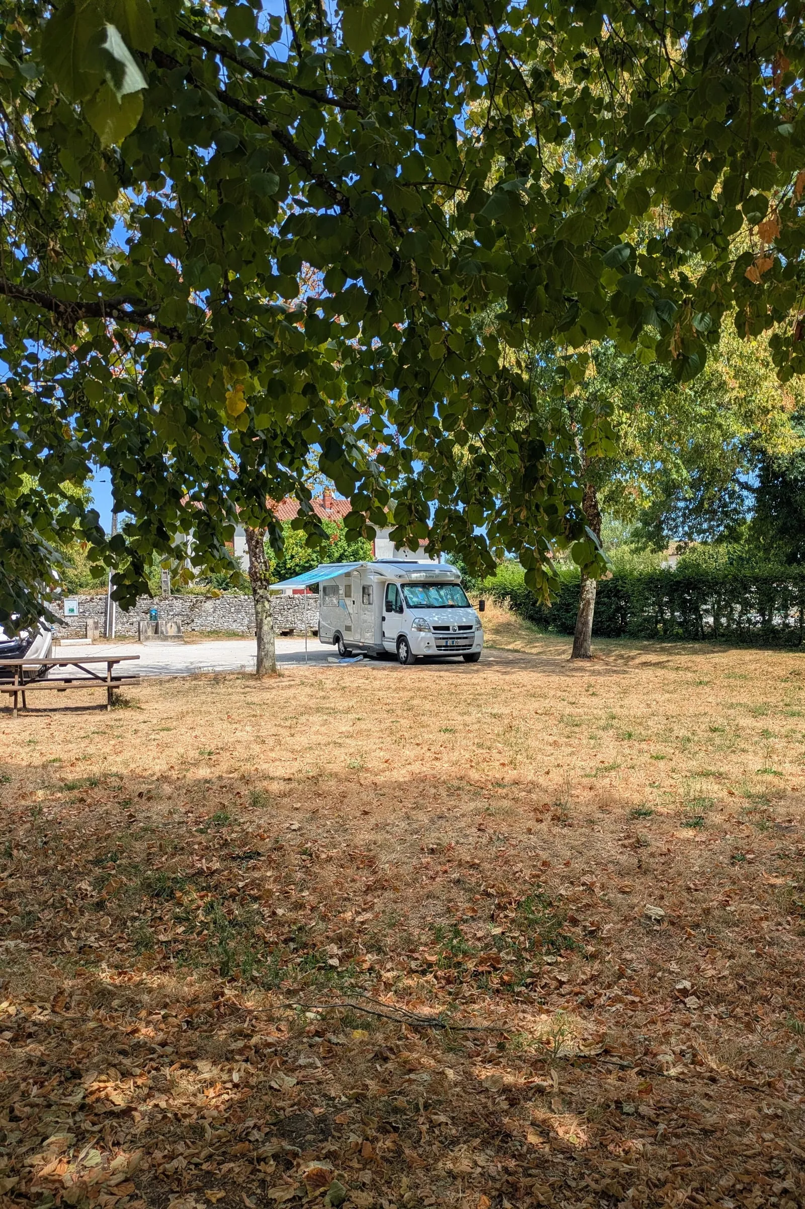 Ember parked in the shade of big lime trees at the Verteuil motorhome aire, awning out, with the dry summer grass of the park in front